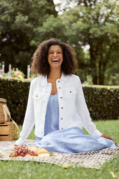 Model wearing Monza Linen Jacket in white layered over a dress, sitting at a picnic scene.