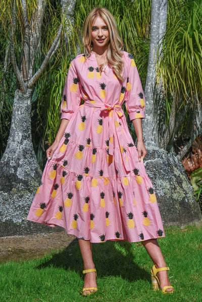 Model wearing the COOP Pineapple Palms Dress in pink, showing the tiered skirt, waist tie, puff sleeves, and all-over pineapple embroidery in a garden setting.