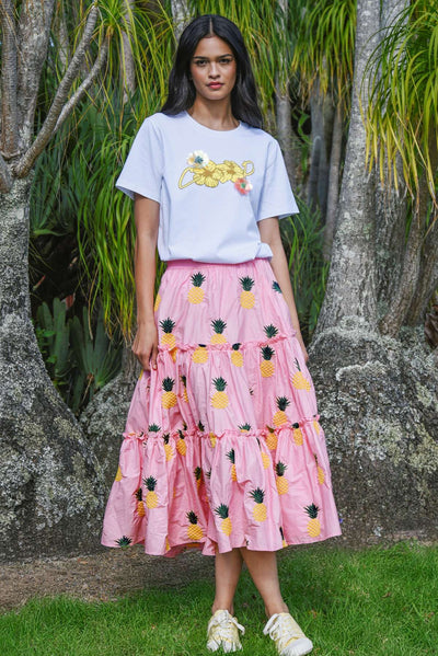 Model wearing the COOP Pineapple Paradise Skirt in pink, featuring tiered ruffles and yellow pineapple embroidery, paired with a white T-shirt in an outdoor setting.