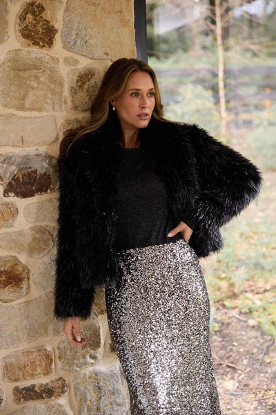 Woman wearing the Brave+True Glamour Fur Jacket Black Sparkle styled with a sequin skirt, standing against a stone wall.